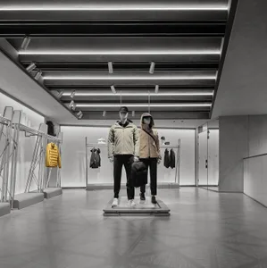 Modern minimalist menswear retail store interior with raw concrete walls, geometric floor tiles, linear LED ceiling lights, and two mannequins dressed in contemporary jackets displayed on a central metal platform.