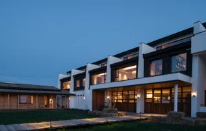 Modern farmhouse exterior at twilight featuring white plaster walls, dark wood cladding, large windows, and a wooden boardwalk leading through a grassy courtyard with hay bales.