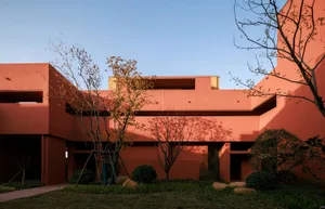 Exterior view of a modern terracotta-colored building with geometric forms, surrounded by a landscaped courtyard with trees and shrubs under a clear blue sky.