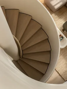 Top-down view of a minimalist spiral staircase made with light oak treads and a smooth white curved central column, adjacent to a woven natural-fiber rug and neutral sofa.