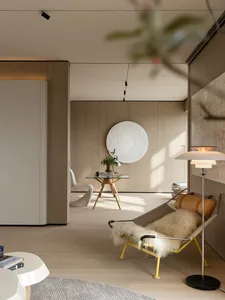 Modern minimalist living room featuring a bright yellow metal lounge chair with sheepskin throw, oak wood paneling, a large circular white textured wall art, and a sculptural white chair in the background.