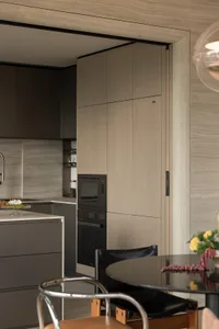 Modern minimalist kitchen featuring taupe wood-grain handleless cabinets, integrated black oven and microwave, marble-look backsplash, and a black leather dining chair in the foreground.