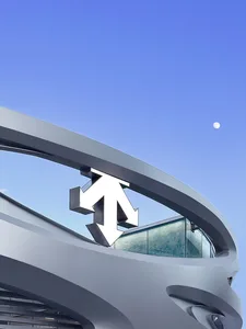 Low angle view of a modern curved metallic building facade featuring a large white and black three-dimensional arrow sculpture against a clear blue evening sky with a visible moon.