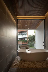 Modern outdoor bathroom featuring a large freestanding terrazzo bathtub on a bed of river pebbles, with dark stone walls and warm wood ceiling overlooking tropical greenery.