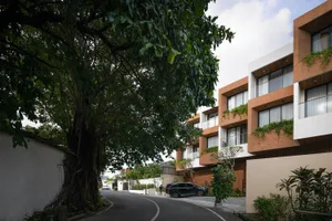 Modern tropical residential building with terracotta panels and white walls, framed by a large leafy tree on a curved street.