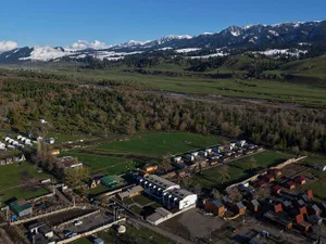 Aerial view of an eco-lodge village featuring modern wooden and white buildings arranged in a valley with snow-capped mountain range in the background.