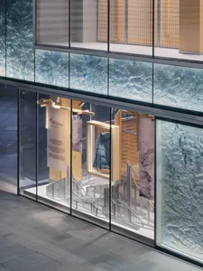 Exterior view of Mountain Down Taipei flagship store at night, showcasing blue textured glass panels, wooden display frames, a hanging blue down jacket, and a sculptural white staircase installation.