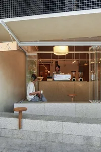 Man sitting on terrazzo steps outside a modern minimalist cafe, enjoying coffee while a barista prepares drinks inside.
