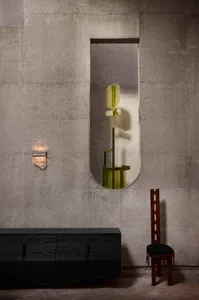 Minimalist brutalist interior featuring raw concrete walls, a black charred wood console, a tall dark wood chair with black cushion, a modern wall sconce, and an olive-green abstract sculpture viewed through a vertical oval opening.