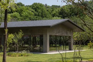Modern concrete house with large glass windows surrounded by green forest and lawn under a clear blue sky.