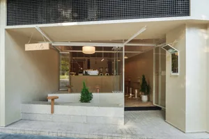 Modern minimalist cafe entrance featuring a raised terrazzo platform with wooden stools, small evergreen trees, and a clean concrete facade with large glass openings.