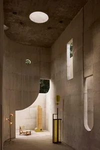 Interior view of a brutalist concrete courtyard house featuring a large circular skylight, curved concrete walls, wooden lounge chair, and yellow-green sculptural lighting elements.