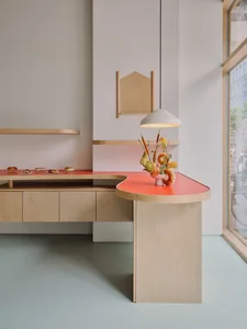 Modern minimalist bakery interior featuring a curved coral-red pastry counter and light birch plywood cabinetry with matching floating shelves, a white pendant lamp, and a floral arrangement with bagels.