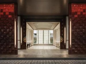 Symmetrical view of a luxurious hotel lobby entrance featuring deep red textured tiled walls, vertical light fixtures, circular art panels with horse motifs, white columns, and a bright courtyard visible in the background.
