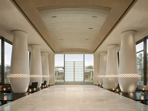 Long symmetrical hotel lobby hallway featuring large white travertine columns with glowing patterned light bands, polished marble floor, and dramatic curved ceiling.