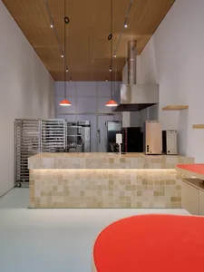 Modern commercial kitchen interior with a large beige ceramic tiled island, warm under-cabinet lighting, two red pendant lamps, wooden ceiling, and stainless steel professional equipment in the background.