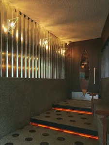 Modern brutalist-style entrance hall with polished terrazzo floor featuring black dots, recessed orange LED step lighting, vertical fluted brass wall with triangular wall sconces, and a dark wood door with a leaf-shaped mirror cutout.