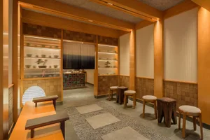 Modern Japanese-style tea room interior with hinoki wood paneling, illuminated display shelves for ceramics, wooden stools, tree-stump seats, stone stepping pads on gravel, and a large paper lantern.