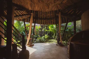 Interior view of a round open-air bamboo pavilion with curved wood ceiling, concrete floor, and lush tropical garden visible all around.