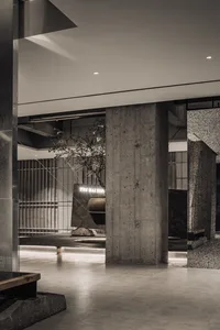 Modern brutalist interior lobby featuring massive raw concrete pillars, terrazzo accents, a large stone bonsai tree, and dramatic architectural lighting.