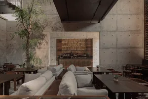 Spacious modern restaurant interior featuring raw concrete walls, light grey modular sofas, dark wood tables, an indoor tree, and a wooden slat bar counter in the background.