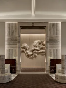 Modern luxury hotel lobby with grey marble pillars, white armchairs, and a large undulating metallic wall sculpture illuminated by warm backlighting.