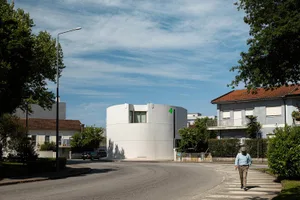 A modern white cylindrical building with minimal windows located at a street corner in a European residential neighborhood, with a man crossing the street in the foreground.