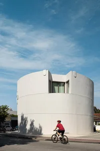 A man in a red shirt riding a bicycle in front of a modern curved white concrete building on a sunny day.