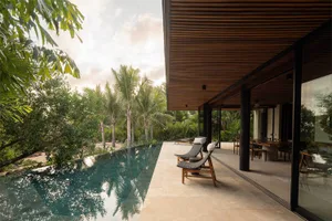 Modern tropical villa with infinity pool, teak lounge chairs on a stone terrace, and lush palm trees in the background under a soft sky.