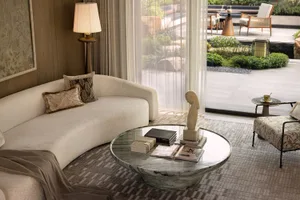 Modern living room featuring a curved cream bouclé sofa, round green marble coffee table with books and sculpture, floor lamp, and large glass doors opening to a landscaped garden.
