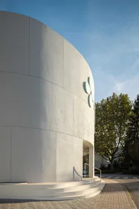 Exterior view of a modern curved white building with smooth stucco finish, sculptural flower logo, wide steps, and metal handrail under a clear blue sky.