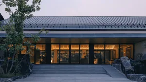 Modern single-story residence with floor-to-ceiling glass facade, concrete steps, and large natural stone landscaping at twilight.