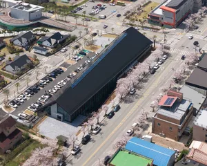 Aerial view of a long modern building with a striking black roof and extensive glass facade, surrounded by cherry blossom trees in full bloom, with parking areas and urban streets visible.