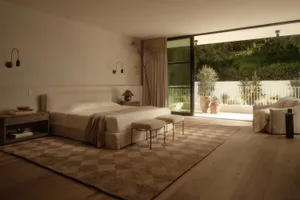 A serene minimalist bedroom featuring a low platform bed with beige linen bedding, a large checkerboard pattern area rug in earthy tones, wooden nightstands, wall-mounted reading lamps, and a large sliding glass door opening to a sunny balcony with potted plants and greenery.