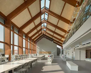 Interior view of a modern two-story library with a high wooden vaulted ceiling, large skylights, long white study tables, and extensive bookshelves lining both floors.