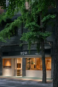 Street view of Shanwei Rice Shop with dark textured concrete upper facade, warm wooden interior visible through large windows, and green ginkgo trees in foreground at night.
