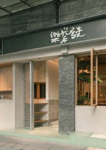 Modern minimalist cafe entrance featuring a tall stacked stone pillar, sage green steps, light wood accents, and large glass openings with plants inside.