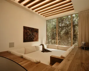 Modern minimalist sunken living room with white sectional sofa, oak wood floor and platform, wooden slat ceiling with lighting, large windows overlooking forest, and abstract painting on the wall.