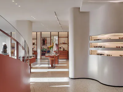Modern minimalist retail boutique interior with curved white walls, terrazzo flooring, wooden display fixtures, and a prominent wall of illuminated shelves displaying sunglasses.