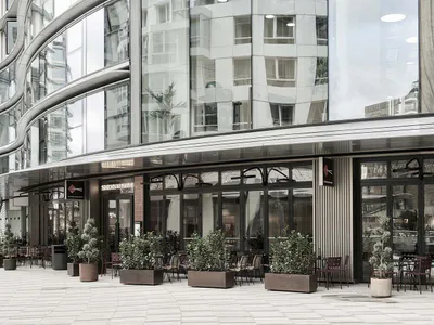 Modern building with curved glass facade and outdoor restaurant seating area featuring potted plants and metal furniture on a paved plaza.