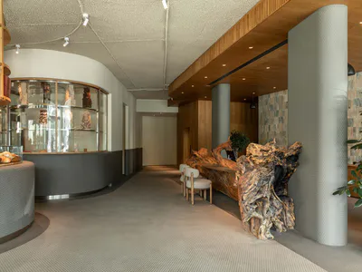 現代麵包店室內，設有弧形灰色蜂巢紋吧台、玻璃甜點展示櫃、擺放紅盒的木製層架，以及大型原木根雕桌與布面座椅。