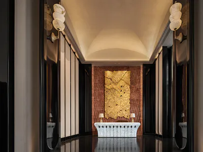 Symmetrical luxury hotel hallway featuring a large textured gold leaf artwork on a copper-toned wall, a white onyx marble console desk with two lamps, dark reflective walls, and sculptural alabaster pendant lights under a curved ceiling.