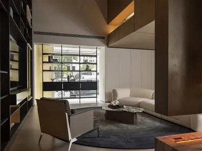 Modern double-height living room featuring dark walnut wood paneling, a curved off-white sofa, a sculptural armchair, and a tall built-in bookshelf wall with warm ambient lighting.