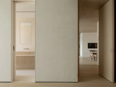 Modern minimalist interior hallway with light oak flooring, white walls, wooden ceiling panels, and open doorways revealing a bathroom and kitchen dining area.