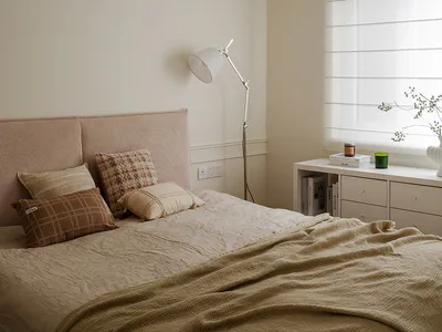 Cozy neutral bedroom with a large textured pinkish headboard, layered beige and brown pillows, a mustard linen throw, sculptural white pendant light, and a white console table under a window with roman blinds.