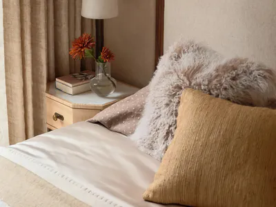 Close-up of a serene bedroom corner featuring a upholstered beige headboard, mustard corduroy pillow, fluffy alpaca wool cushion, marble pendant lamp, and a wooden nightstand with marble top holding a glass vase with orange flowers.