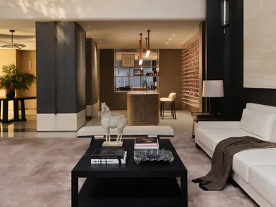 Modern luxury double-height living room with travertine stone walls, white sectional sofa, black coffee table, and upper mezzanine level overlooking the space.