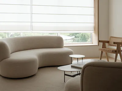 Modern minimalist living room featuring a large curved beige boucle sofa, marble nesting tables, a wooden cane chair, and a striking triple-ring modern chandelier hanging above a large window with sheer roller blinds.