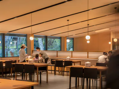 Modern minimalist family restaurant interior featuring light wood tables, black chairs, spherical pendant lights, and large windows overlooking a forest.
