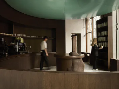 Modern café interior featuring a curved dark terrazzo service counter, matching central table with organic-shaped stools, sage green curved ceiling, and warm wood shelving under natural window light.